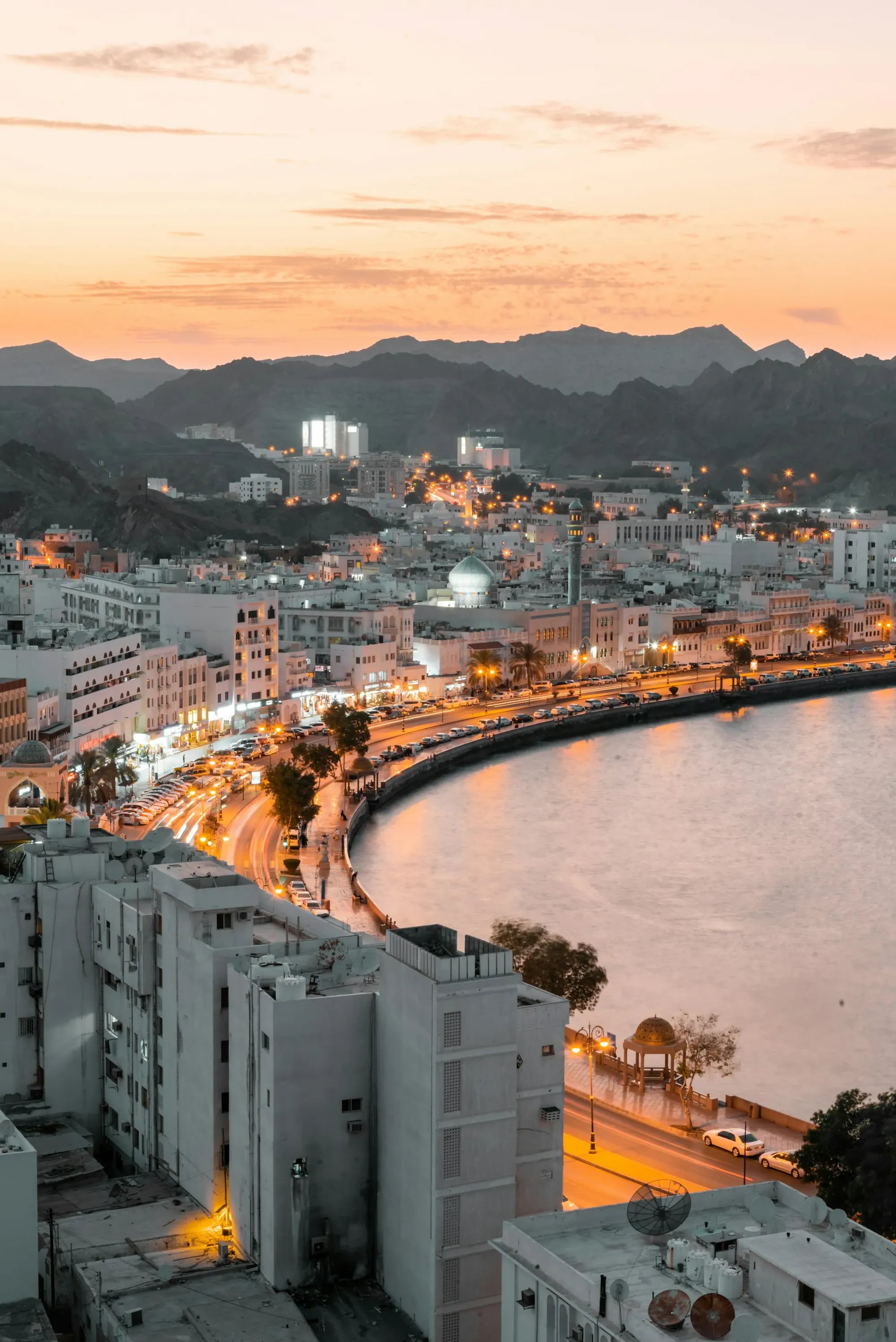 Muscat's waterfront curving along the bay at dusk, with mountains behind