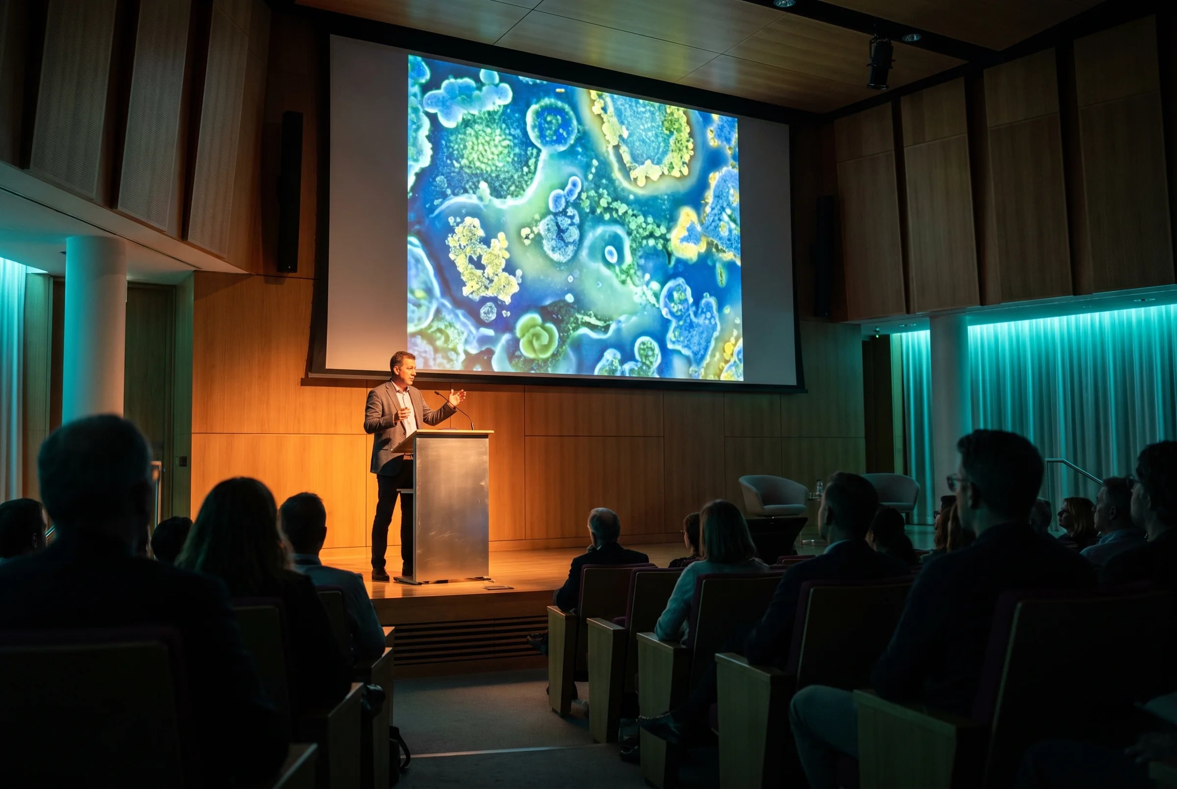 A speaker at a lectern in front of a large screen showing colourful bacterial colonies, with the audience in silhouette